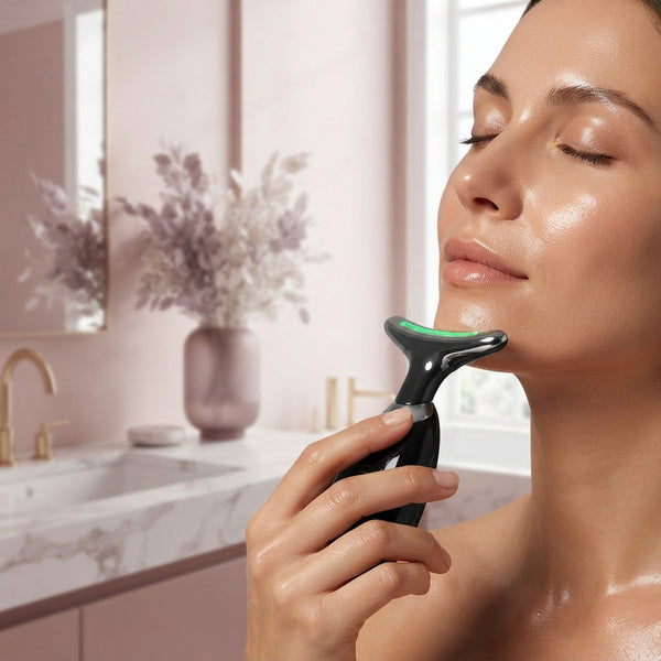 Woman using a handheld facial massager along her jawline in a modern bathroom with soft pink accents.